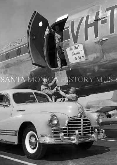 Man standing in a plane receiving air mail from two men below.